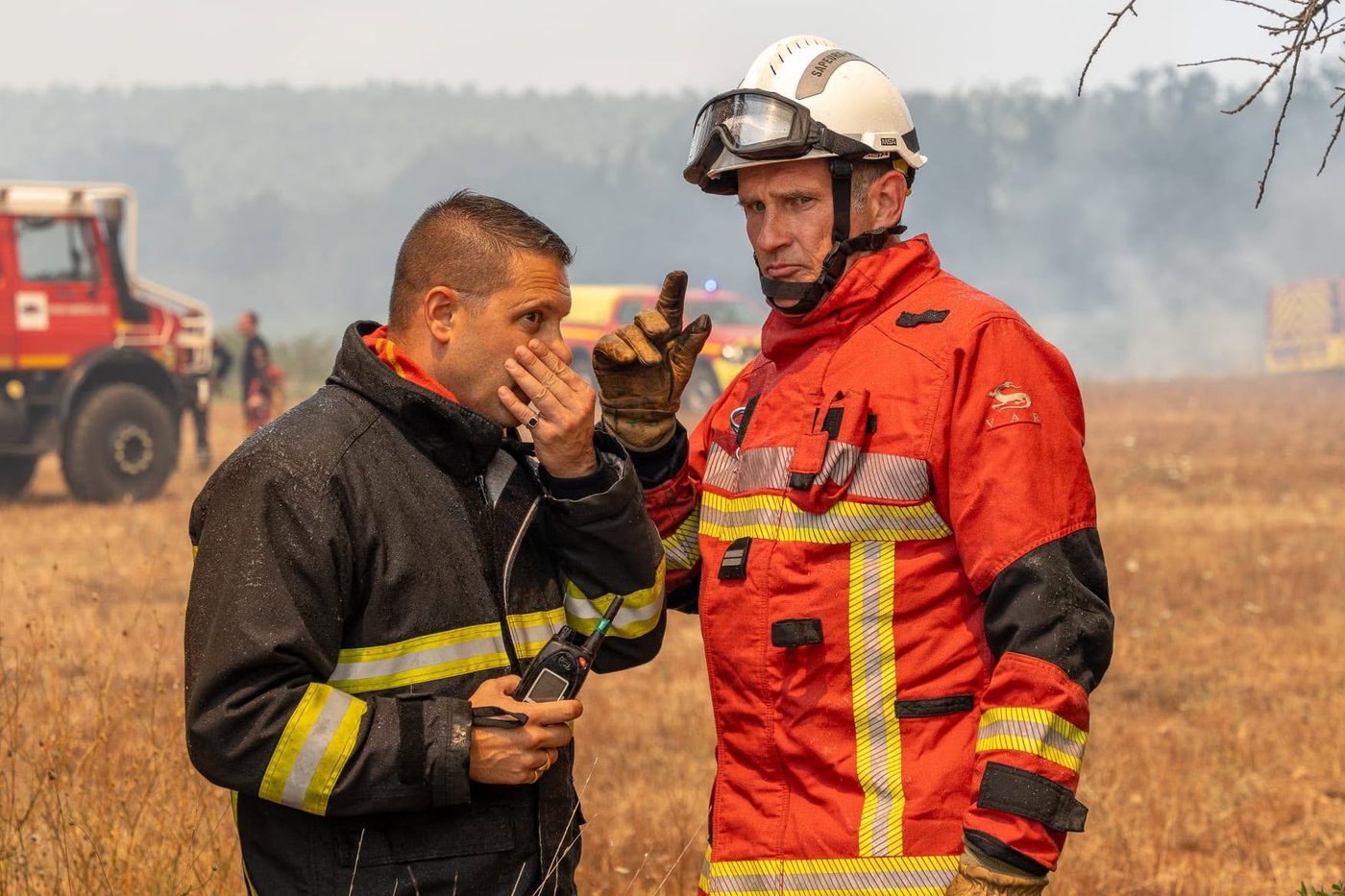 Een officier van de Sapeurs-Pompiers du Var overlegt aan de rand van een brandgebied, met op de achtergrond een blusvoertuig en rook boven een droge vlakte.