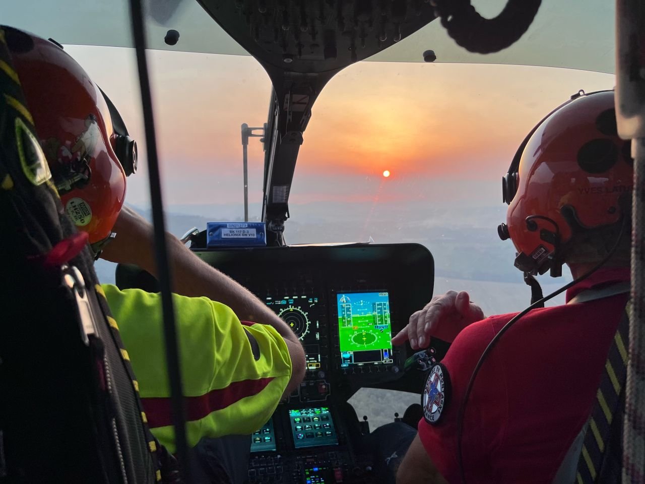 Twee bemanningsleden van een reddingshelikopter tijdens een verkenningsvlucht boven het brandgebied in de Var, met de ondergaande zon op de achtergrond.